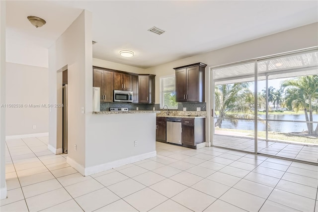 kitchen with light stone countertops, a water view, backsplash, stainless steel appliances, and dark brown cabinetry