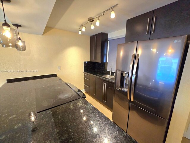 kitchen featuring stainless steel refrigerator with ice dispenser, dark brown cabinetry, sink, decorative light fixtures, and dark stone counters