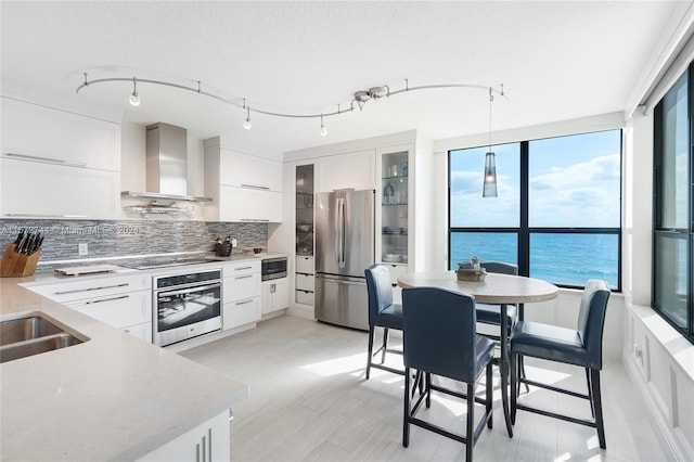 kitchen with appliances with stainless steel finishes, wall chimney exhaust hood, backsplash, white cabinetry, and hanging light fixtures