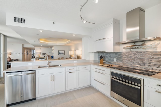 kitchen with wall chimney exhaust hood, kitchen peninsula, appliances with stainless steel finishes, and a tray ceiling