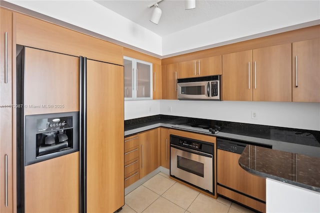 kitchen featuring appliances with stainless steel finishes, brown cabinetry, glass insert cabinets, light tile patterned flooring, and dark stone counters