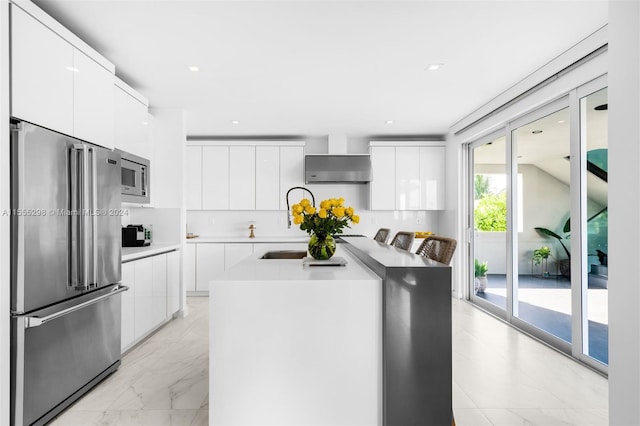 kitchen featuring white cabinetry, wall chimney range hood, stainless steel appliances, light tile floors, and a center island with sink