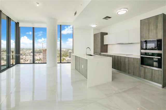 kitchen with white cabinets, appliances with stainless steel finishes, light tile floors, a wall of windows, and an island with sink