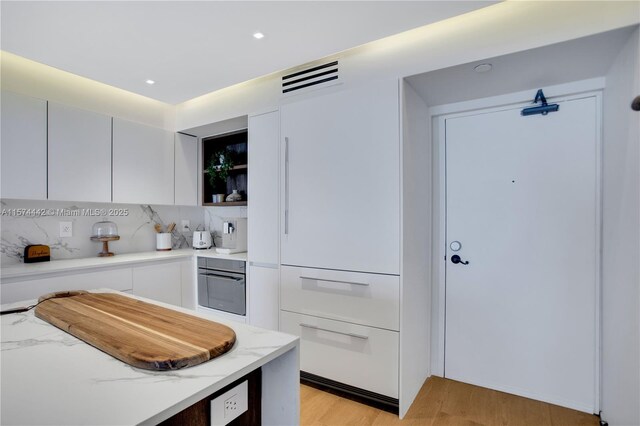 kitchen with light stone counters, white cabinetry, light wood-type flooring, backsplash, and oven