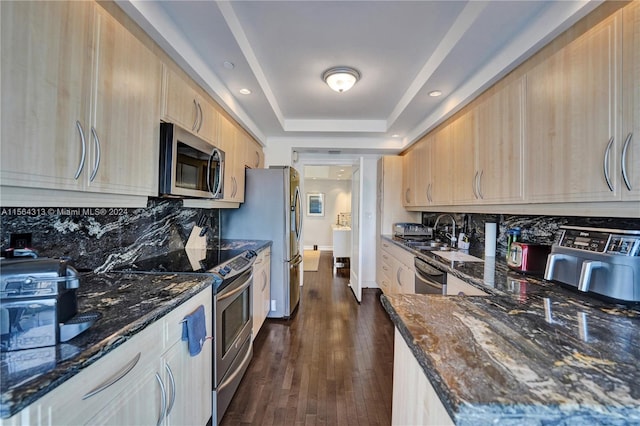 kitchen featuring appliances with stainless steel finishes, dark hardwood / wood-style floors, dark stone counters, light brown cabinetry, and a raised ceiling