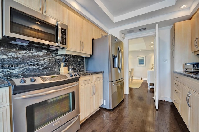 kitchen with appliances with stainless steel finishes, light brown cabinets, a raised ceiling, dark stone counters, and dark hardwood / wood-style floors