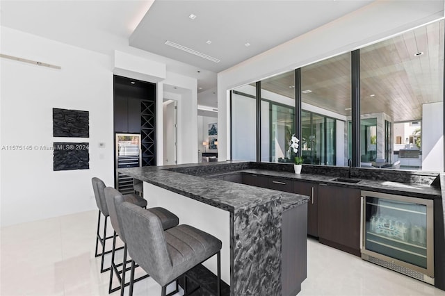 kitchen with beverage cooler, light tile floors, dark brown cabinets, dark stone countertops, and a kitchen breakfast bar