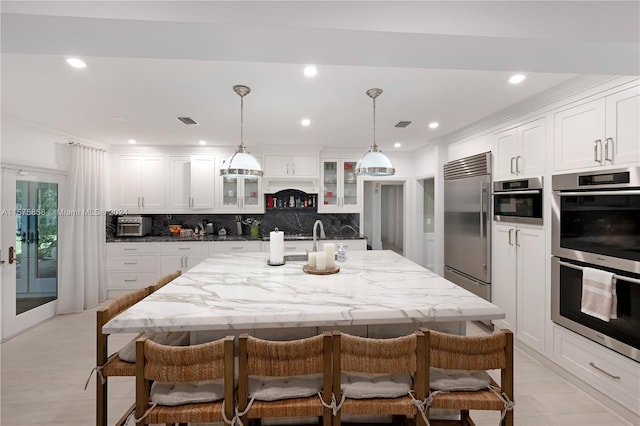 kitchen featuring appliances with stainless steel finishes, backsplash, white cabinetry, a center island with sink, and pendant lighting