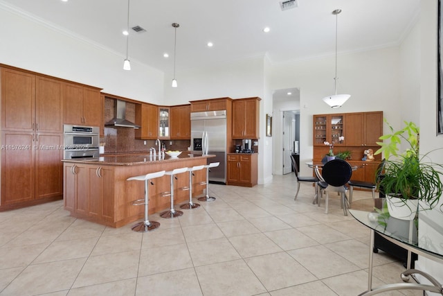 kitchen featuring a high ceiling, wall chimney range hood, an island with sink, a kitchen bar, and stainless steel appliances