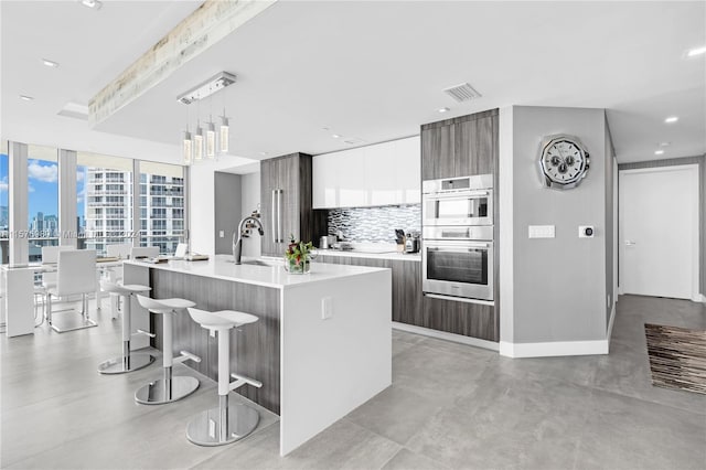 kitchen featuring hanging light fixtures, double oven, tasteful backsplash, a kitchen island with sink, and white cabinets