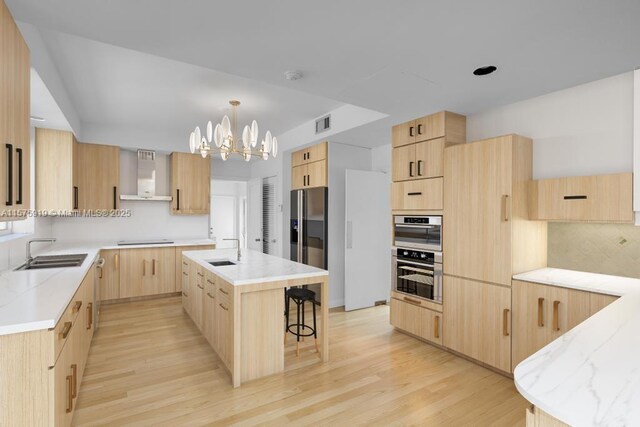 kitchen with light brown cabinetry, sink, wall chimney range hood, and an island with sink