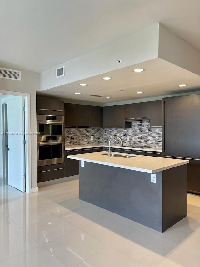 kitchen with double oven, dark brown cabinets, sink, tasteful backsplash, and an island with sink