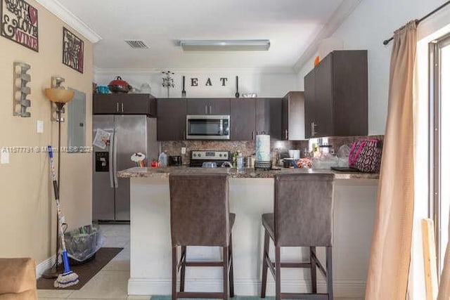 kitchen featuring light tile patterned flooring, dark brown cabinets, decorative backsplash, appliances with stainless steel finishes, and a kitchen breakfast bar