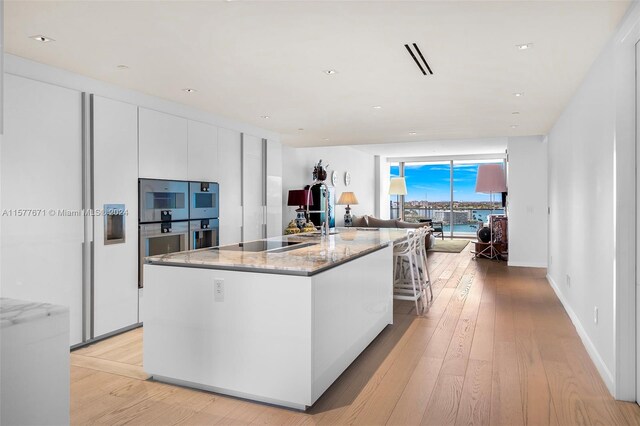 kitchen with white cabinets, light wood-type flooring, black electric cooktop, a center island with sink, and light stone countertops