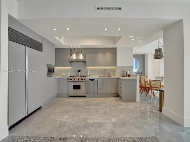 kitchen with wall chimney exhaust hood, gray cabinetry, a tray ceiling, kitchen peninsula, and built in appliances