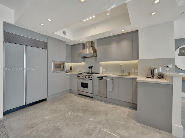 kitchen featuring built in appliances, a raised ceiling, sink, and wall chimney exhaust hood