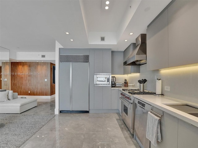 kitchen featuring light tile flooring, wall chimney range hood, a raised ceiling, gray cabinetry, and built in appliances