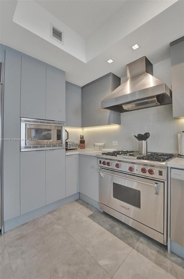 kitchen with appliances with stainless steel finishes, wall chimney exhaust hood, light tile flooring, gray cabinetry, and a tray ceiling