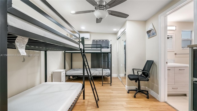 bedroom with a wall mounted air conditioner, light wood-type flooring, ceiling fan, and connected bathroom