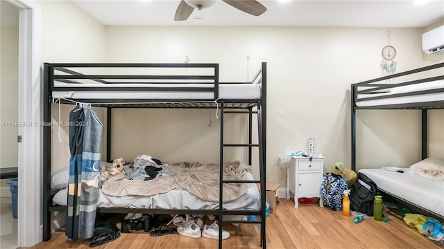 bedroom with a wall unit AC, wood-type flooring, and ceiling fan