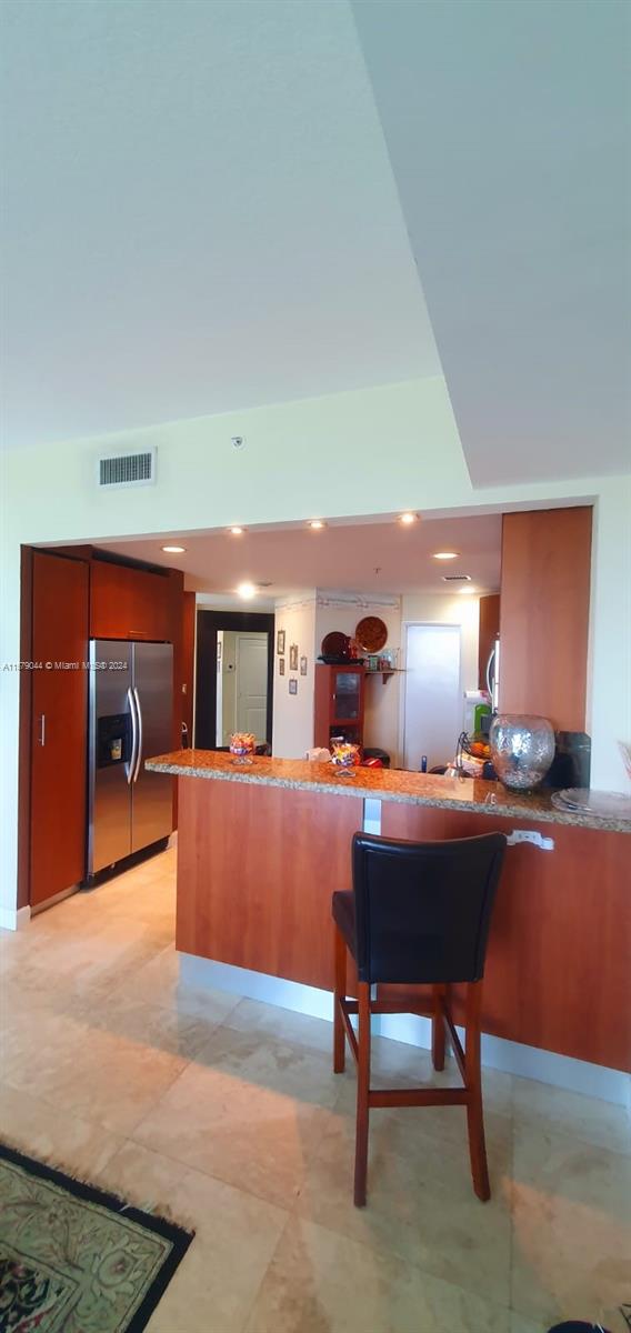 kitchen featuring a kitchen bar, light tile flooring, stainless steel fridge with ice dispenser, kitchen peninsula, and light stone countertops