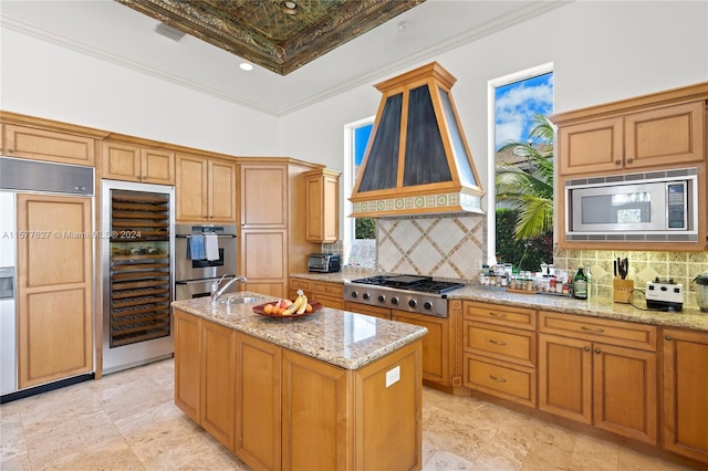 kitchen featuring built in appliances, custom exhaust hood, a kitchen island with sink, light tile flooring, and tasteful backsplash