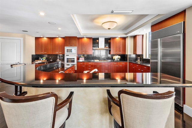 kitchen featuring visible vents, dark countertops, a breakfast bar, a tray ceiling, and wall chimney range hood