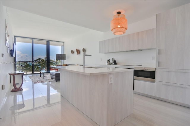 kitchen with oven, black electric stovetop, light brown cabinetry, a wall of windows, and an island with sink