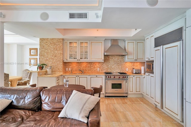 kitchen with built in appliances, a tray ceiling, backsplash, wall chimney range hood, and sink