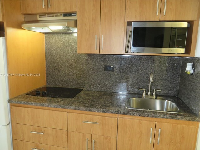 kitchen with wall chimney range hood, dark stone countertops, black electric cooktop, sink, and tasteful backsplash