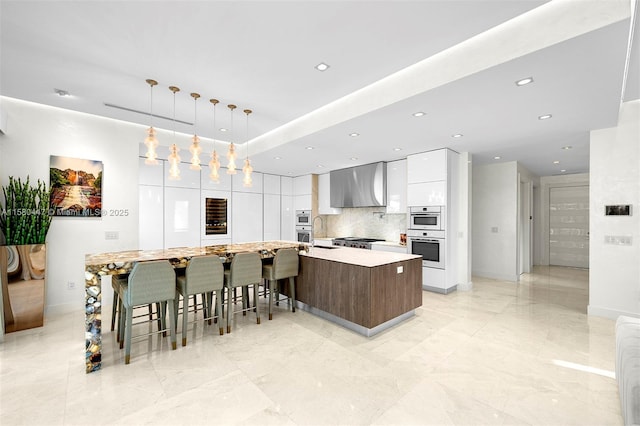 kitchen with white double oven, wall chimney range hood, a large island with sink, white cabinets, and hanging light fixtures