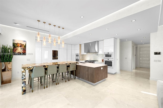 kitchen with double oven, modern cabinets, a spacious island, wall chimney exhaust hood, and white cabinetry