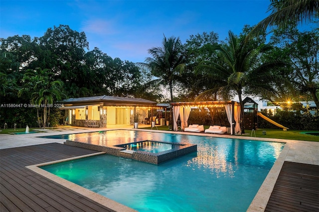 pool at dusk with an in ground hot tub, a playground, and a deck