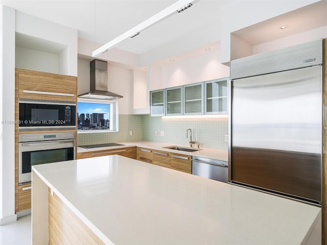 kitchen with wall chimney range hood, tasteful backsplash, sink, built in appliances, and a center island