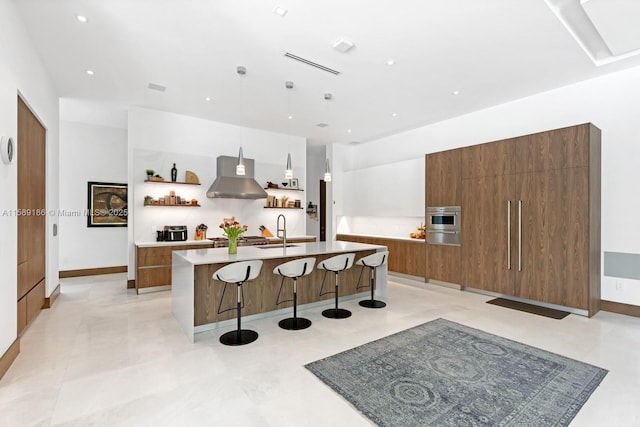 kitchen with sink, wall chimney exhaust hood, hanging light fixtures, a kitchen breakfast bar, and a center island with sink