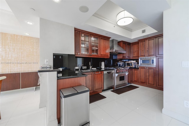 kitchen with appliances with stainless steel finishes, kitchen peninsula, light tile patterned flooring, a raised ceiling, and wall chimney exhaust hood