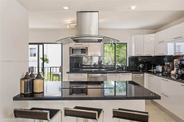 kitchen featuring stainless steel appliances, white cabinetry, island exhaust hood, a kitchen breakfast bar, and tasteful backsplash