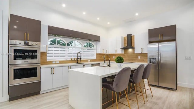 kitchen with a breakfast bar, wall chimney range hood, sink, an island with sink, and appliances with stainless steel finishes