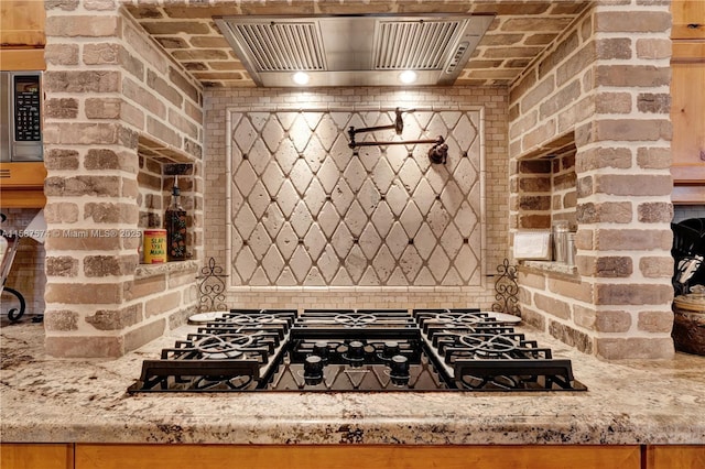 kitchen with stainless steel microwave, premium range hood, brick wall, light stone counters, and black gas cooktop