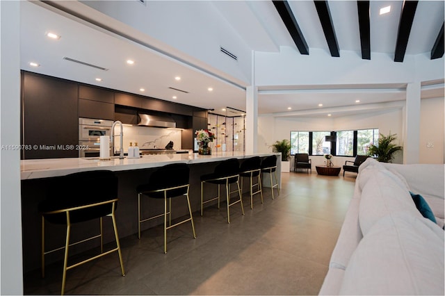 kitchen featuring wall chimney range hood, a kitchen breakfast bar, tile floors, beam ceiling, and stainless steel double oven