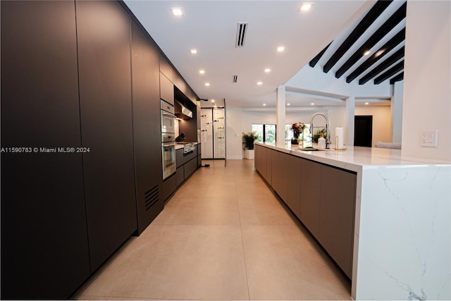 kitchen with light stone countertops, beamed ceiling, double oven, sink, and light tile flooring