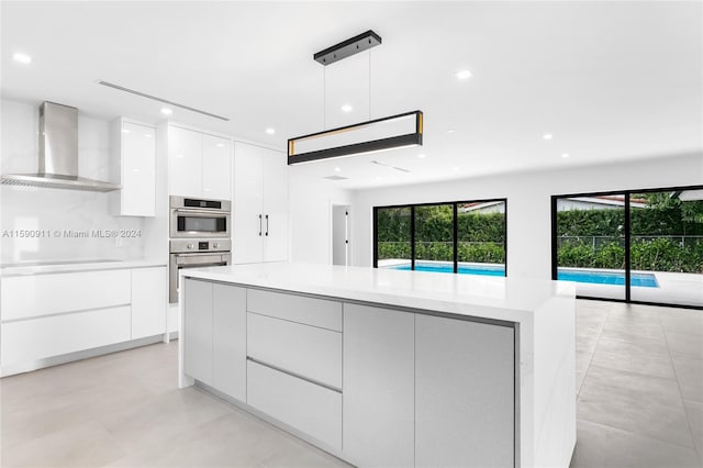 kitchen with a center island, wall chimney exhaust hood, hanging light fixtures, stainless steel double oven, and white cabinetry