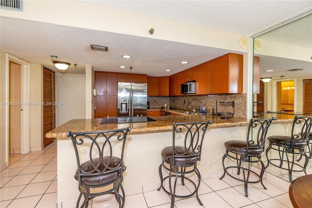 kitchen with a textured ceiling, appliances with stainless steel finishes, kitchen peninsula, backsplash, and light tile floors