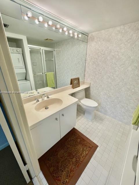 bathroom featuring toilet, vanity, tile patterned floors, stacked washing maching and dryer, and a shower with door