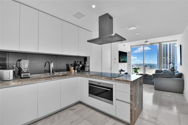 kitchen featuring island range hood, light tile patterned floors, stainless steel oven, black electric cooktop, and white cabinetry