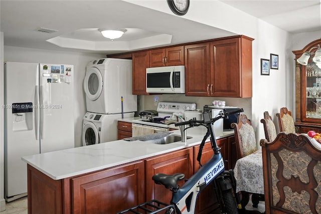 kitchen featuring light stone countertops, a tray ceiling, kitchen peninsula, white appliances, and stacked washer / drying machine