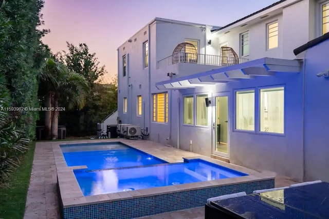 back house at dusk featuring a balcony and a swimming pool with hot tub