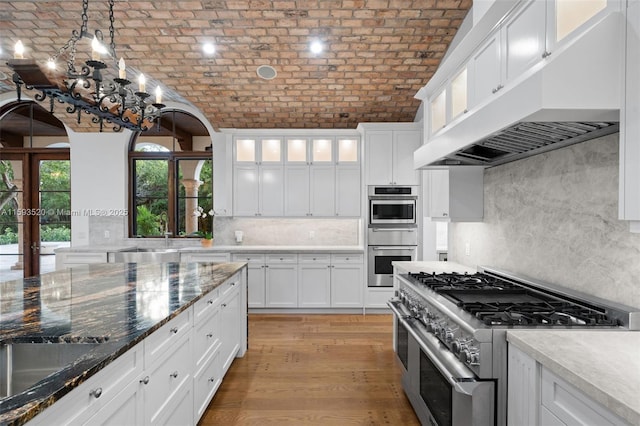 kitchen featuring appliances with stainless steel finishes, wall chimney exhaust hood, white cabinets, and brick ceiling