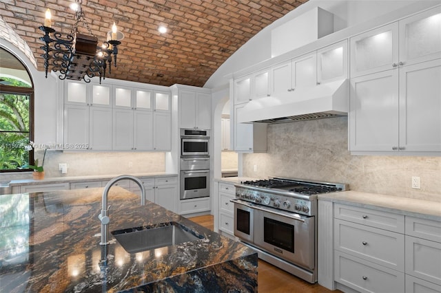 kitchen featuring brick ceiling, stainless steel appliances, sink, dark stone countertops, and white cabinets