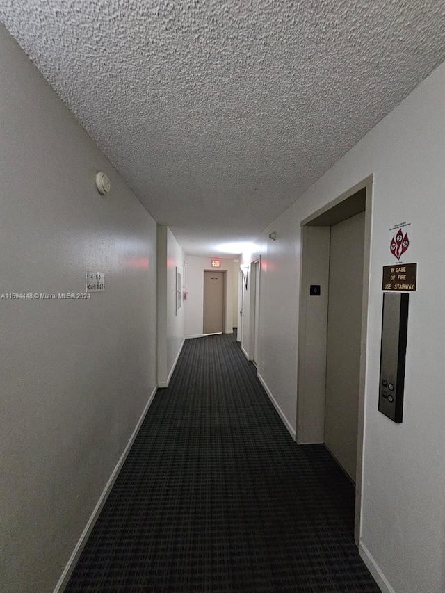 corridor featuring dark colored carpet, a textured ceiling, elevator, and baseboards